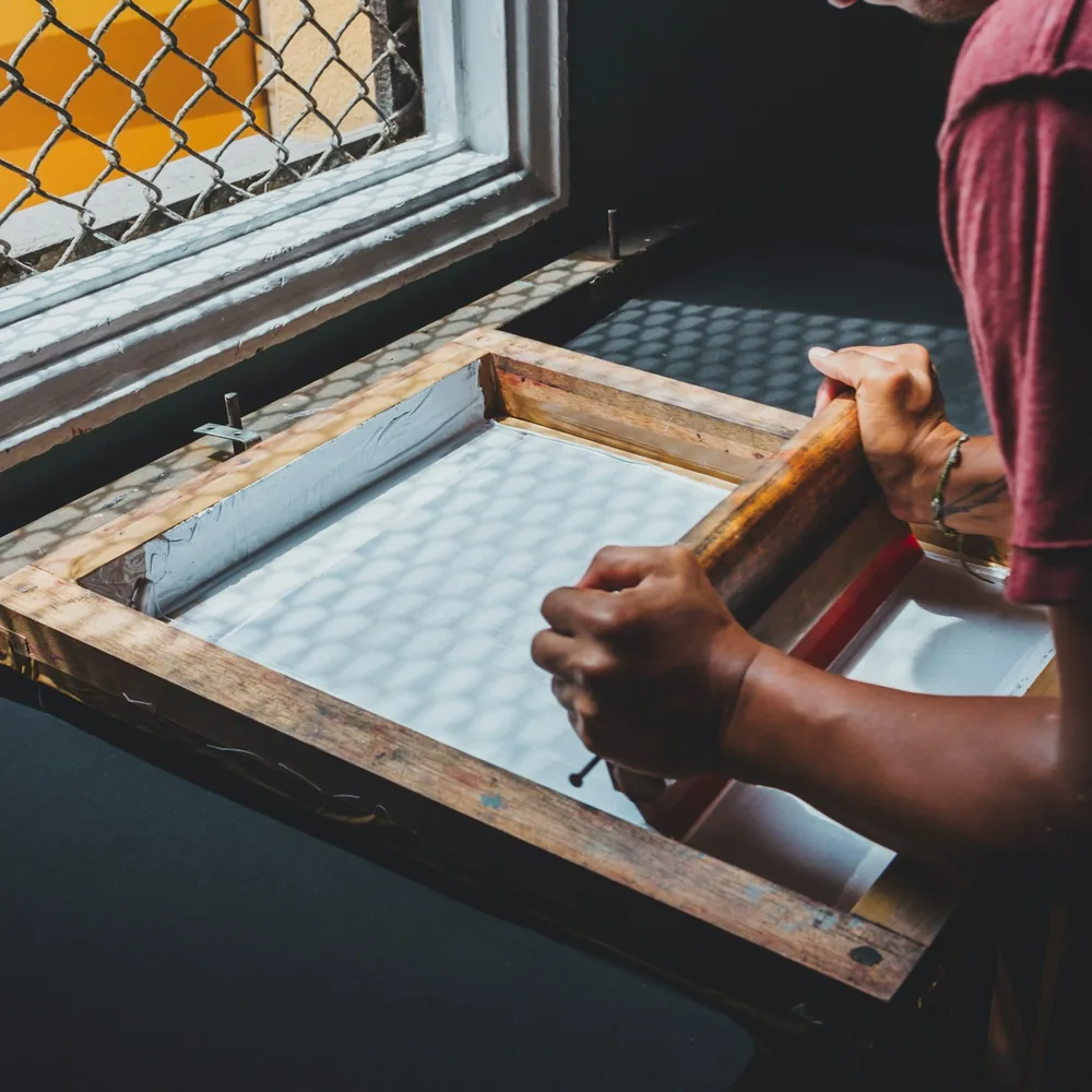 Manos sujetando una racleta sobre un bastidor de madera con la malla tensada, junto a la ventana de un taller de serigrafía